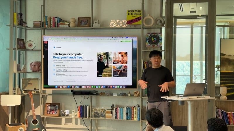 Khai presenting Audon at 500 Global, beside a screen showing the Audon page, "Talk to your computer. Keep your hands free."